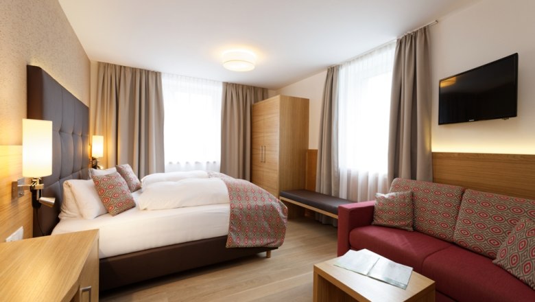 Modern hotel room with double bed, sofa, wardrobe and TV.