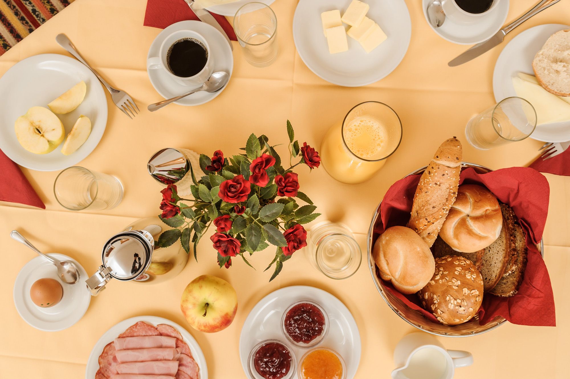 Ein gedeckter Frühstückstisch mit Brötchen, Aufschnitt, Marmelade, Kaffee, Orangensaft und einem Blumenstrauß in der Mitte.