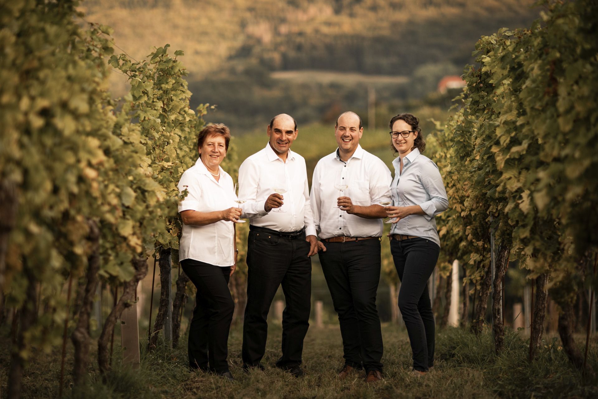 Vier Personen stehen lächelnd in einem Weinberg mit Weingläsern in der Hand.
