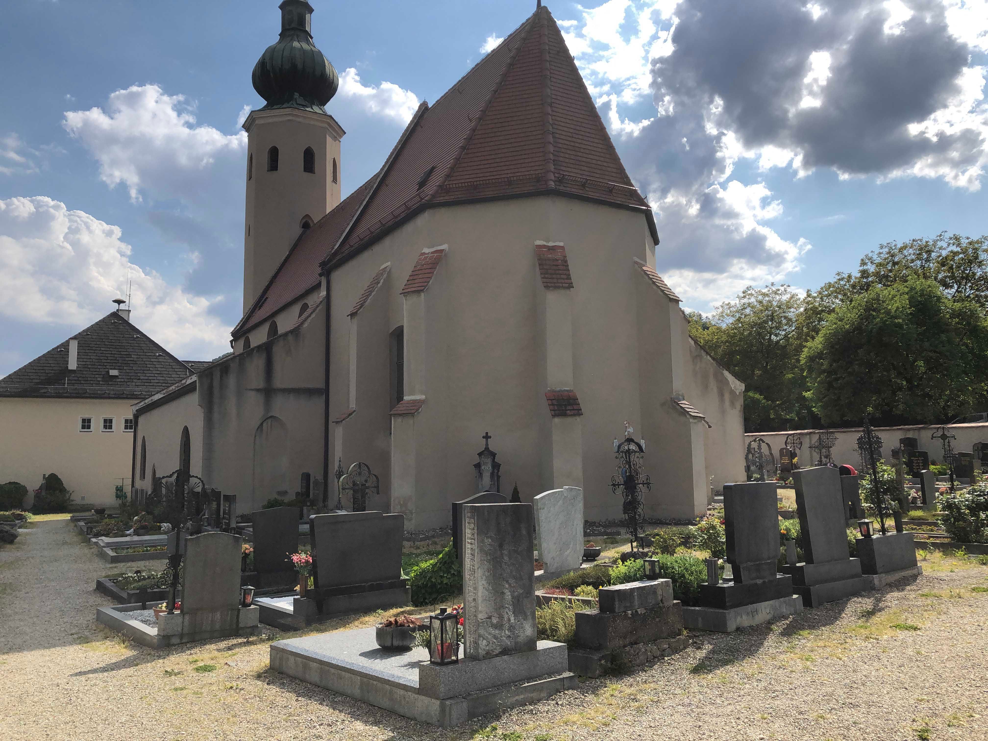 Kirche mit Friedhof und Grabsteinen im Vordergrund, bewölkter Himmel.