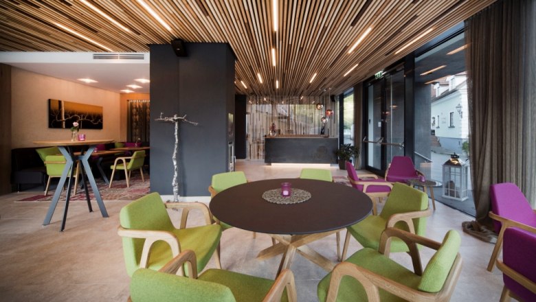 Interior view of a modern hotel lounge with wooden elements, green and purple chairs and a round table.