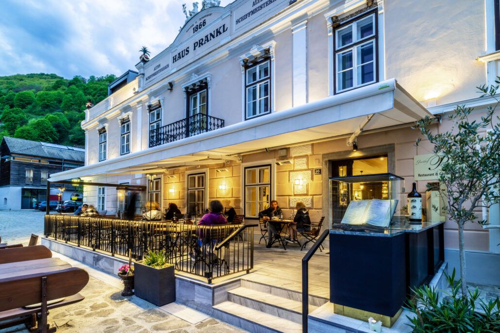 Außenansicht des Gasthofs Prankl mit beleuchteter Terrasse und Gästen an Tischen.