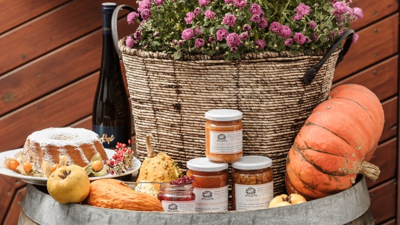 Ein Arrangement aus hausgemachten Spezialitäten mit Kürbis, Marmelade, Kuchen und Blumen in einem Korb auf einem Fass.