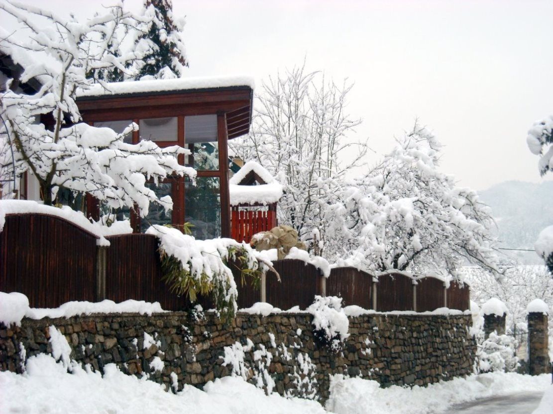 Ein verschneites Haus mit Holzzaun und Steinmauer, umgeben von schneebedeckten Bäumen.