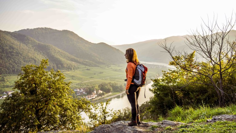 Eine Frau mit Rucksack steht auf einem Hügel und blickt auf eine Flusslandschaft mit Bergen im Hintergrund.