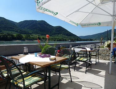 Ein Gastgarten mit Tischen und Stühlen unter Sonnenschirmen, mit Blick auf die Donau und bewaldete Hügel im Hintergrund.