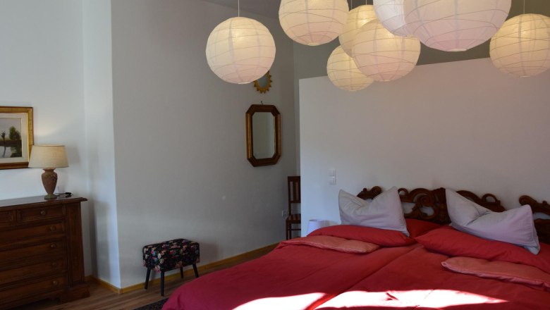 Bedroom with double bed, red comforters, wooden chest of drawers, lamp, mirror and paper lamps on the ceiling.