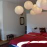 Bedroom with double bed, red comforters, wooden chest of drawers, lamp, mirror and paper lamps on the ceiling.