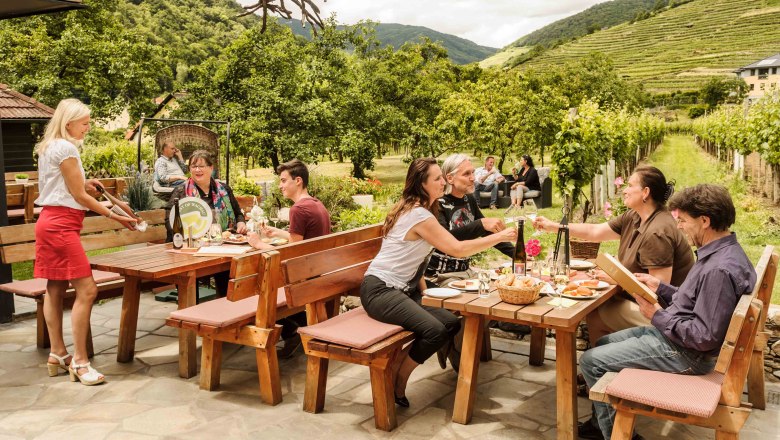 Menschen sitzen an Holztischen im Freien, umgeben von Weinbergen und Bäumen, während eine Kellnerin Getränke serviert.
