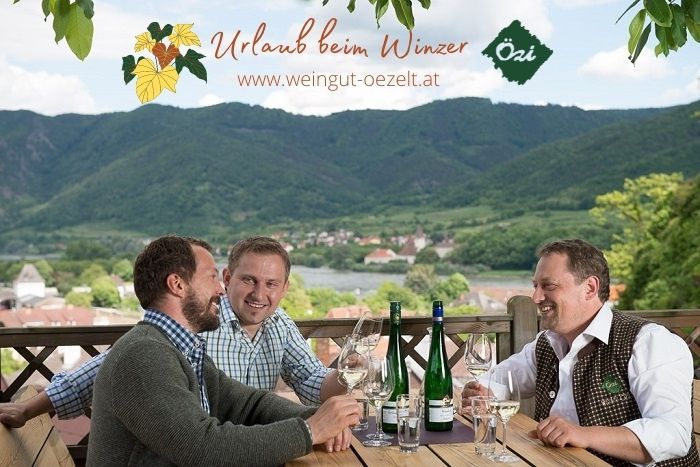 Drei Männer sitzen an einem Tisch mit Weinflaschen und Gläsern, im Hintergrund eine malerische Landschaft mit Hügeln.