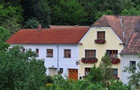 A traditional house with red roof tiles and flower boxes in front of the windows, surrounded by green vegetation.