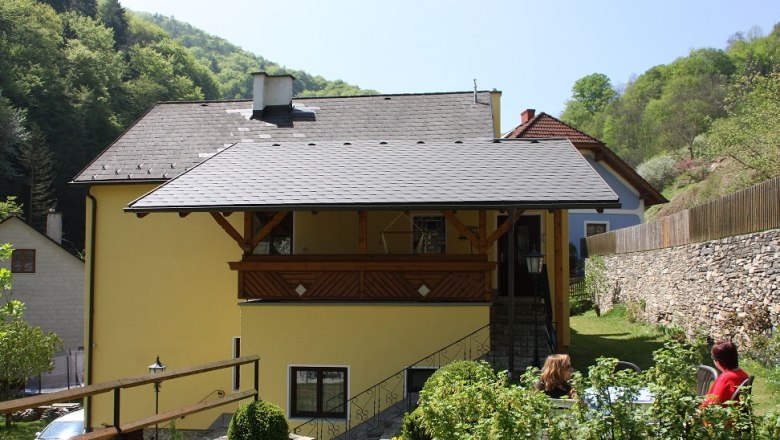 Gelbes Haus mit Balkon, umgeben von grüner Landschaft und Steinmauer.