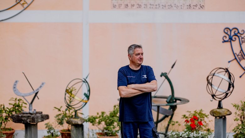 Ein Mann steht mit verschränkten Armen vor einer Wand mit Sonnenuhren und Pflanzen.