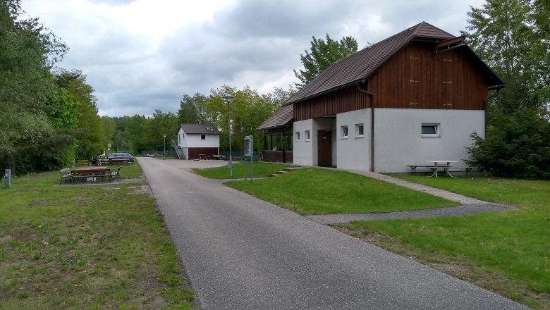 A path leads past a white building with wooden cladding, surrounded by trees and meadows, under a cloudy sky.