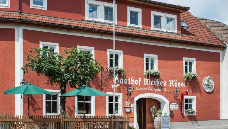 Rotes Gebäude mit weißen Fensterrahmen und grünen Sonnenschirmen vor dem Eingang.