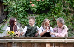 Four people in traditional dress clink glasses of wine outside in front of a wall covered in plants.