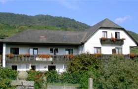 Ein zweistöckiges Haus mit Balkon und Blumen, umgeben von grüner Landschaft und Hügeln im Hintergrund.
