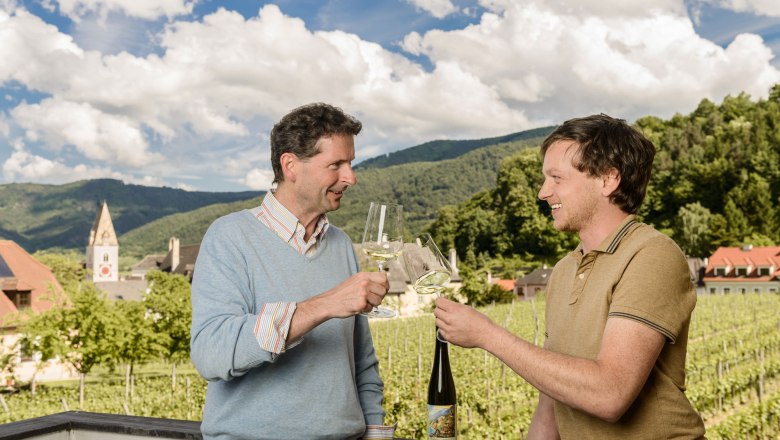 Zwei M&auml;nner sto&szlig;en mit Weingl&auml;sern an, im Hintergrund Weinberge und ein Kirchturm.