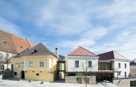 Zwei Geb&auml;ude mit gelber und wei&szlig;er Fassade, verbunden durch einen modernen Glasgang, unter blauem Himmel.