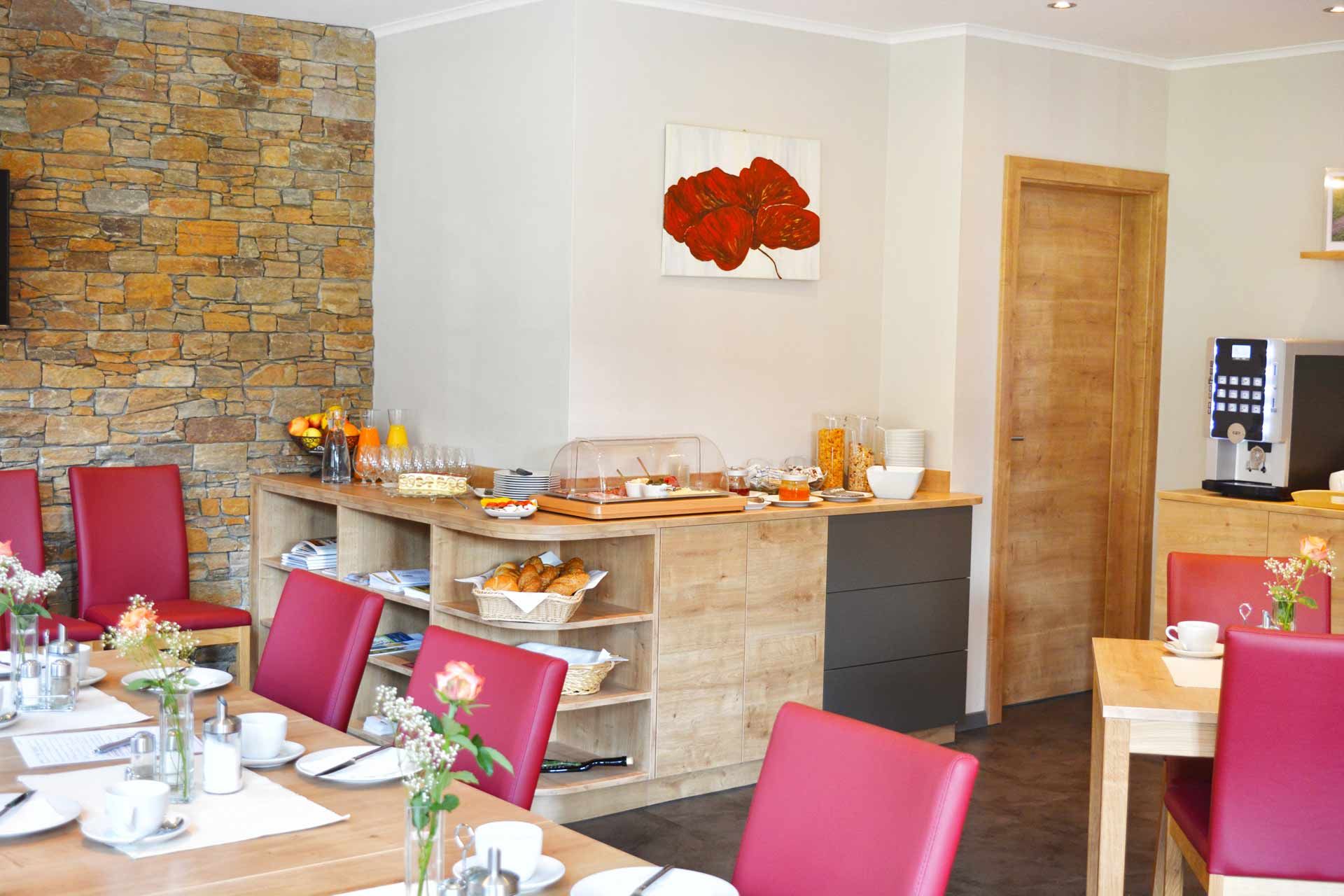 Ein Frühstücksbuffet in einem gemütlichen Raum mit roten Stühlen, Holzmöbeln und einer Steinwand. Auf dem Buffet sind Croissants, Müsli und Orangensaft.