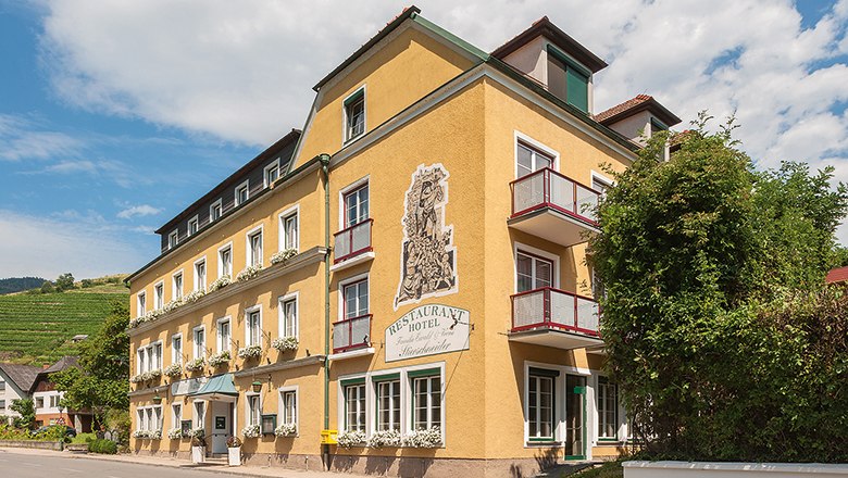 Stierschneider's Weinhotel Wachau, © Johann Perger Yellow hotel building with balconies and plants, blue sky in the background.