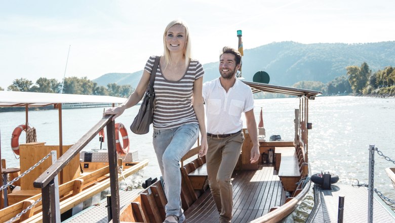 A man and a woman step off a boat onto a jetty. A river and wooded hills can be seen in the background.