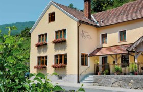 Au&szlig;enansicht eines gelben G&auml;stehauses mit roten Dachziegeln und Blumenk&auml;sten an den Fenstern.