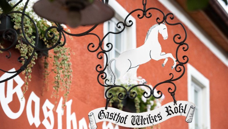 Metallschild mit weißem Pferd und Schriftzug 'Gasthof Weißes Rößl' vor rotem Gebäude.