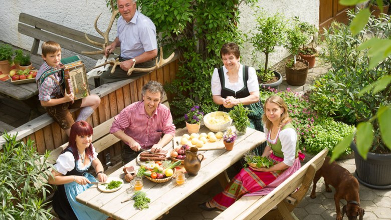 Eine Familie in traditioneller Kleidung sitzt an einem Tisch im Garten und bereitet eine Mahlzeit vor.