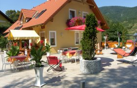 Guest house with terrace, tables, chairs and parasols, surrounded by plants and mountains in the background.