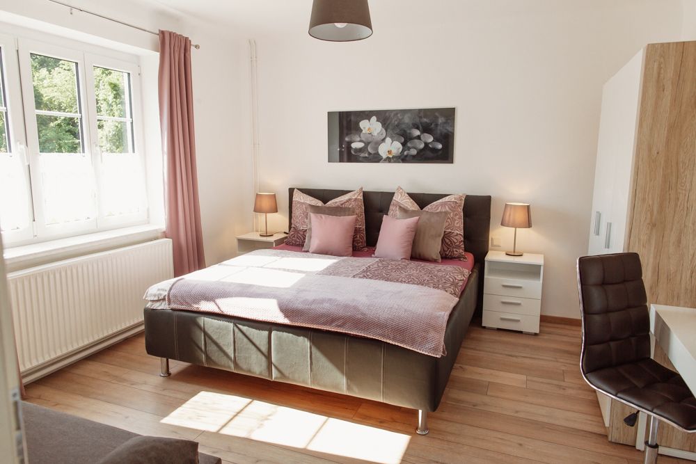 Helles Schlafzimmer mit Doppelbett, zwei Nachttischen und einem Fenster mit rosa Vorhängen.