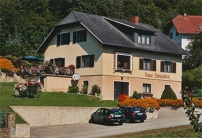 Ein zweistöckiges Haus mit der Aufschrift 'Haus Donaublick', umgeben von Blumen und Bäumen. Zwei Autos parken vor dem Haus.
