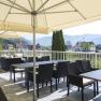 Terrasse mit Tischen und Stühlen unter Sonnenschirmen, Blick auf Landschaft und Häuser.