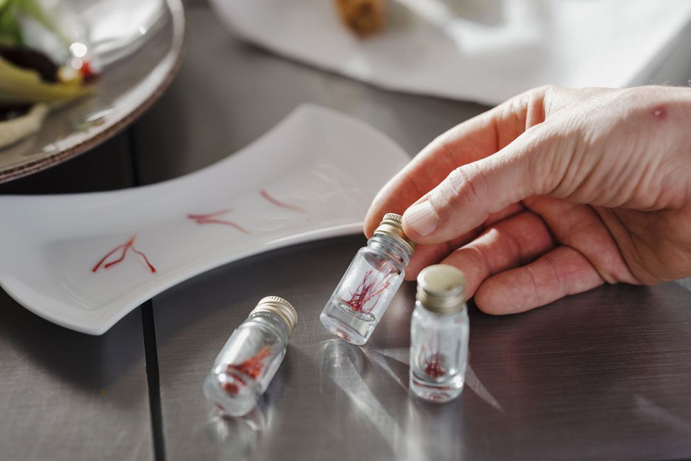 Hand hält ein kleines Glasfläschchen mit Safranfäden, daneben zwei weitere Fläschchen und ein Teller mit Safran.