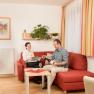 Ein Paar sitzt auf einem roten Sofa in einem gemütlichen Wohnzimmer. Der Mann hält eine Weinflasche, die Frau lächelt. Pflanzen und ein Bild schmücken den Raum.