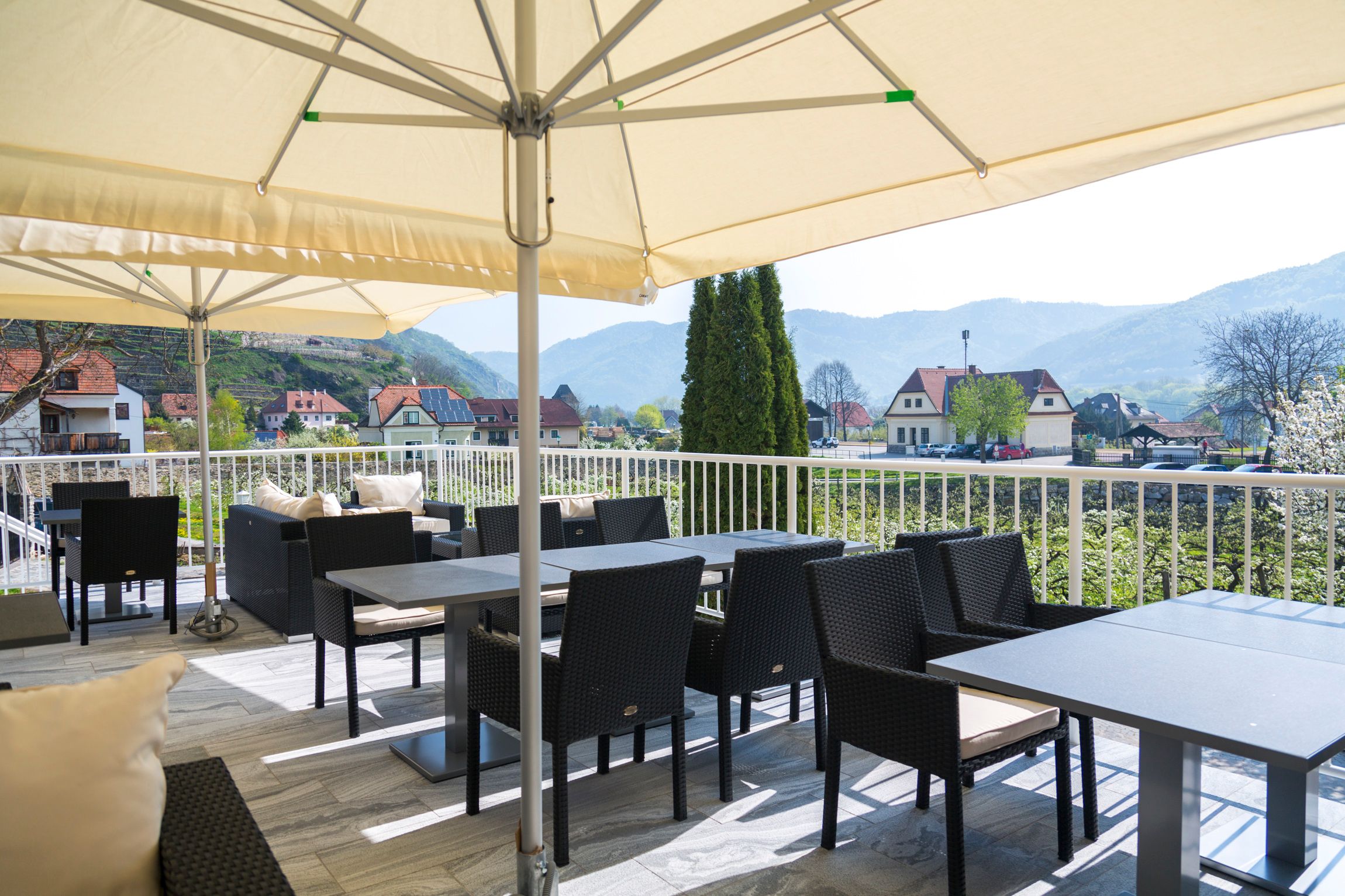 Terrasse mit Tischen und Stühlen unter Sonnenschirmen, Blick auf Landschaft und Häuser.