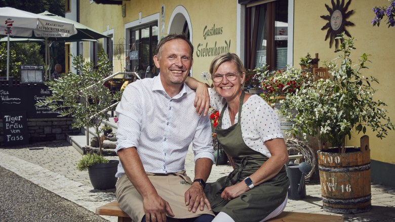 Smiling couple in front of Gasthaus Siebenhandl, surrounded by plants and decoration.