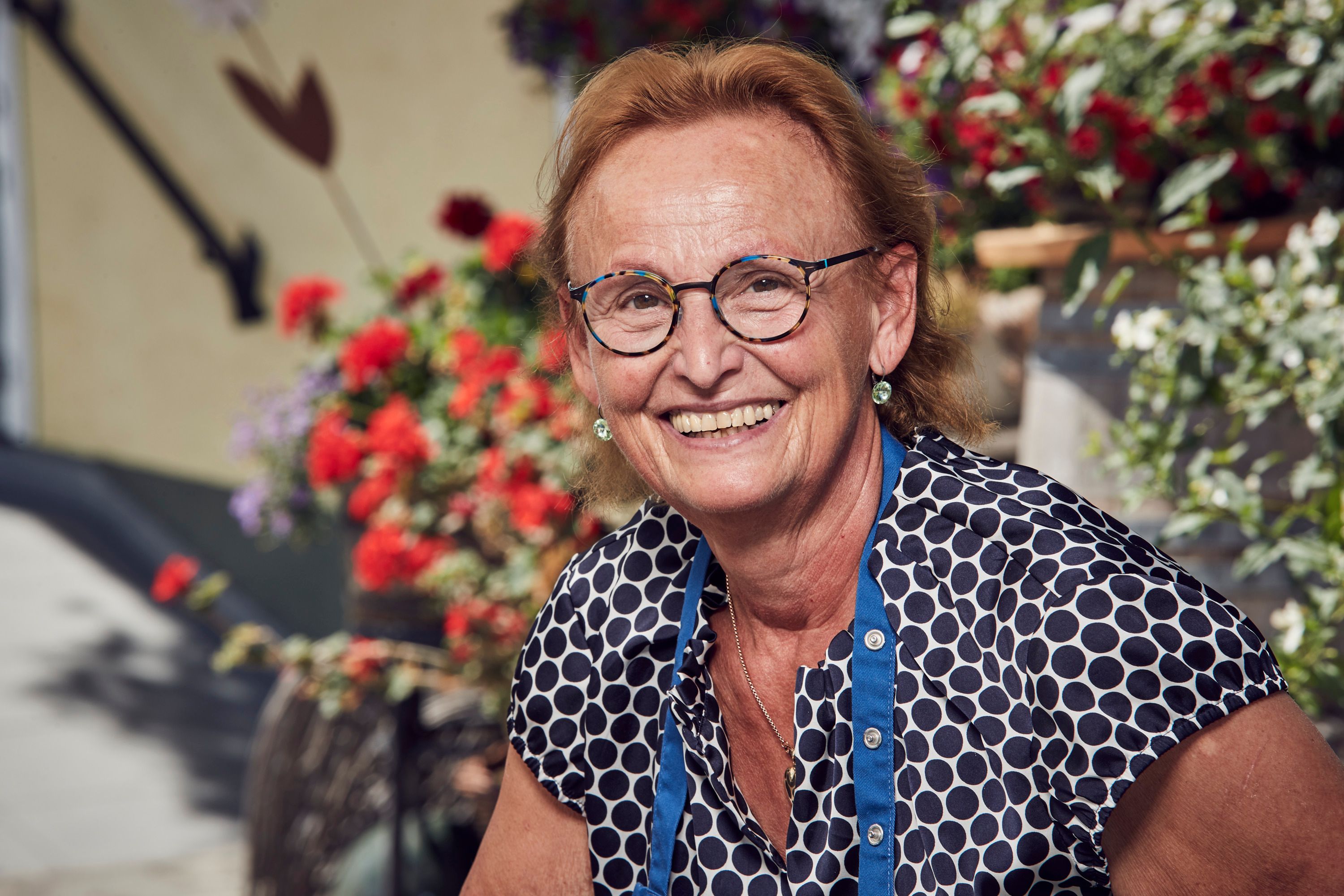 Eine lächelnde Frau mit Brille und gepunkteter Bluse vor blühenden Pflanzen.