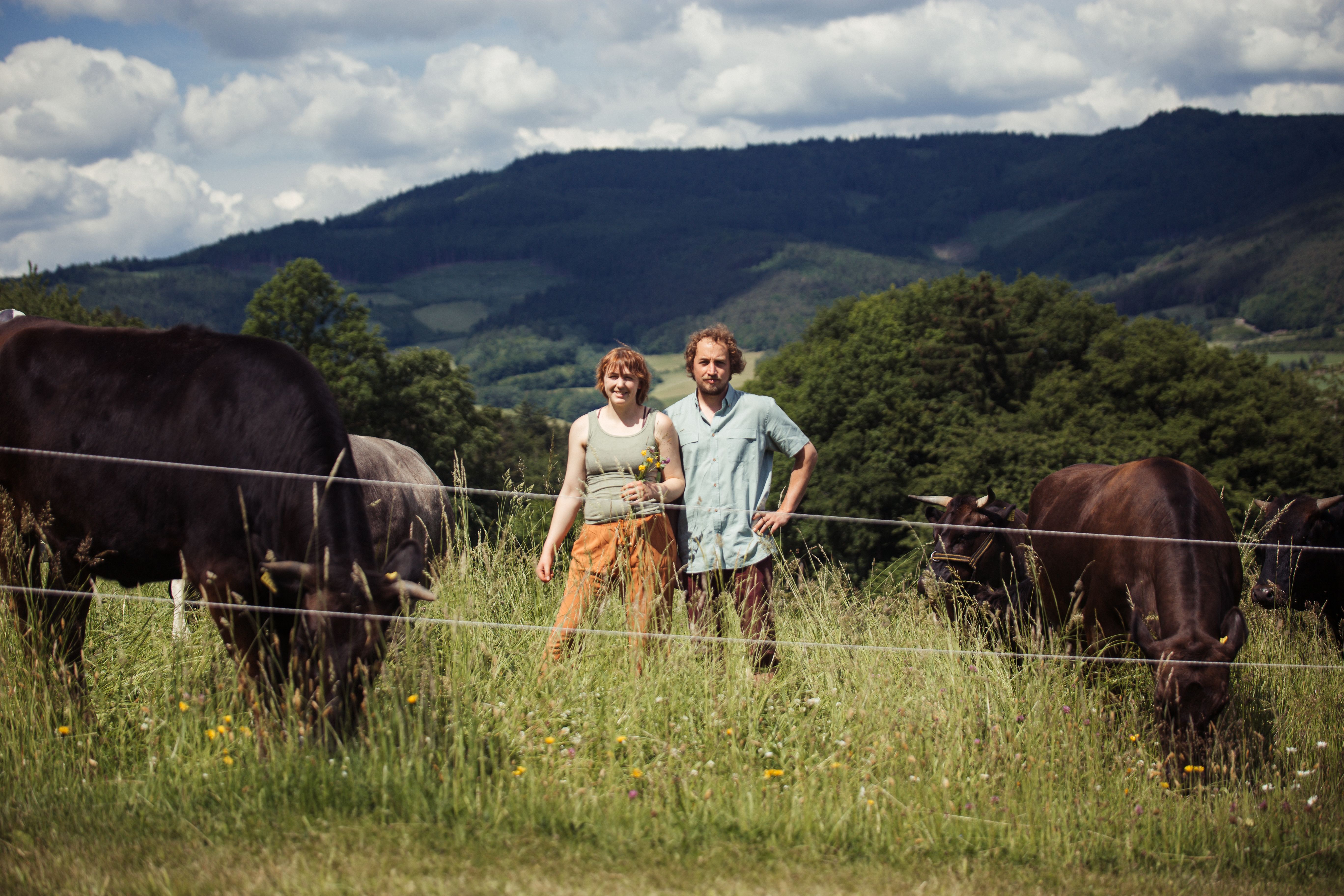 Ein Bauer und seine Frau stehen neben ihren Kühen auf einer eingezäunten Weide. 