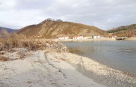A quiet beach with pebbles, a village and wooded hills in the background.