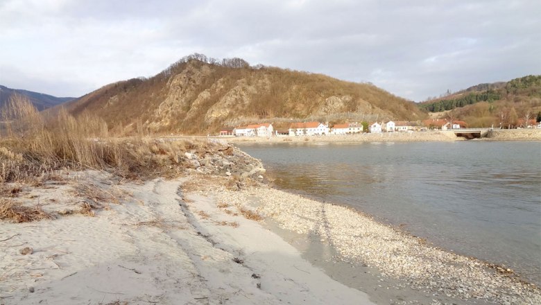 strand, © Kowalski Ein ruhiger Strand mit Kieselsteinen, im Hintergrund ein Dorf und bewaldete Hügel.