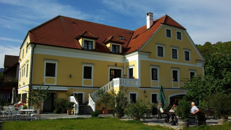 Gelbes Geb&auml;ude mit rotem Dach und Terrasse, umgeben von B&auml;umen.