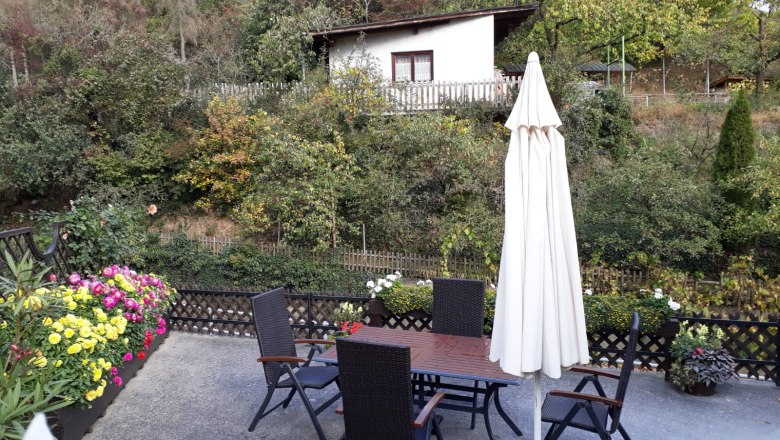 Terrasse mit Tisch, Stühlen und Sonnenschirm, umgeben von Blumen und Bäumen.