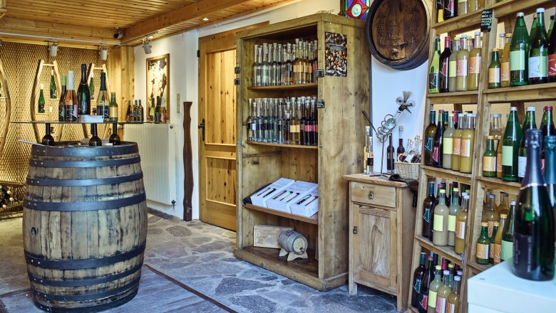 Innenansicht eines Ab-Hof-Ladens mit Holzregalen voller Flaschen und einem Weinfass als Tisch.