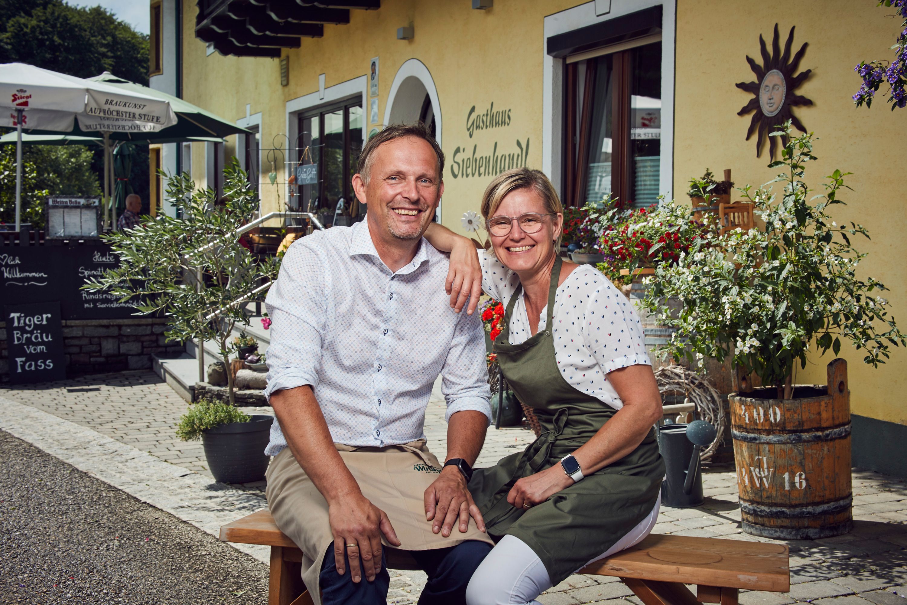 Lächelndes Paar vor Gasthaus Siebenhandl, umgeben von Pflanzen und Dekoration.