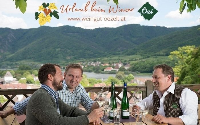 Three men sit at a table with bottles of wine and glasses, a picturesque landscape with hills in the background.