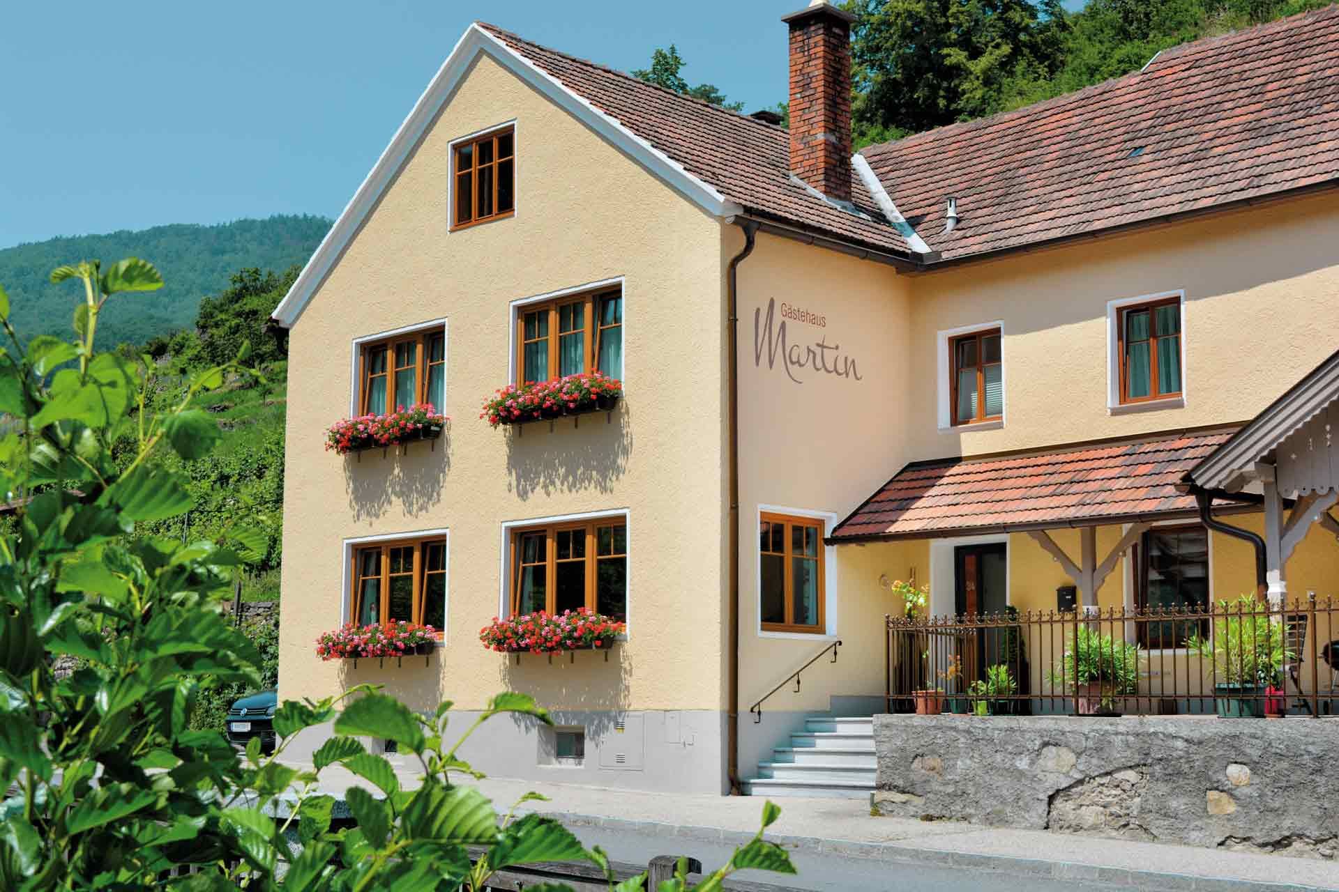 Außenansicht eines gelben Gästehauses mit roten Dachziegeln und Blumenkästen an den Fenstern.
