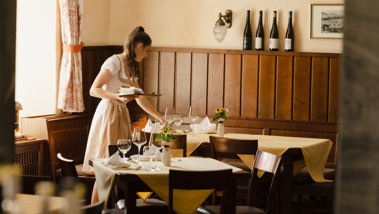 Eine Frau in traditioneller Kleidung deckt einen Tisch in einem gemütlichen Restaurant mit Holzwänden und Weinflaschen im Hintergrund.
