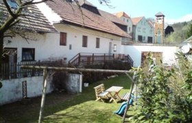 Ein ländlicher Hof mit weißen Gebäuden, Holztisch und Stühlen im Garten, Schaukel und Rutsche im Vordergrund.