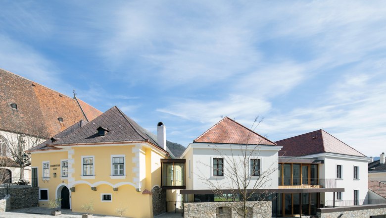 Zwei Gebäude mit gelber und weißer Fassade, verbunden durch einen modernen Glasgang, unter blauem Himmel.